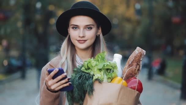 Young beautiful woman wearing stylish coat standing in the autumn park holding package of products and using smartphone. Girl looking into the camera. Shopping, healthy eating, internet-shop concept — Stock Video