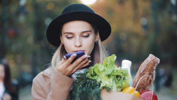 Giovane bella donna che indossa cappotto elegante in piedi nel parco contenente pacchetto di prodotti e utilizzando il riconoscimento vocale smartphone, detta pensieri, messaggio di composizione vocale — Video Stock