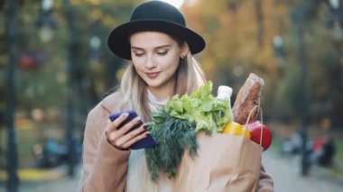Şık ceket ayakta paket ürünlerin tutarak ve smartphone kullanarak sonbahar parkta giyen genç güzel kadın. Alışveriş, sağlıklı beslenme, internet salonu konsept