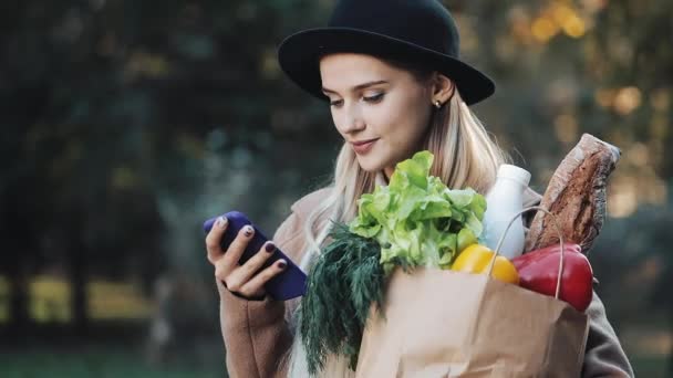 Mujer hermosa joven con un abrigo elegante de pie en el parque de otoño que sostiene el paquete de productos y el uso de teléfonos inteligentes. Compras, alimentación saludable, concepto de tienda en Internet — Vídeo de stock