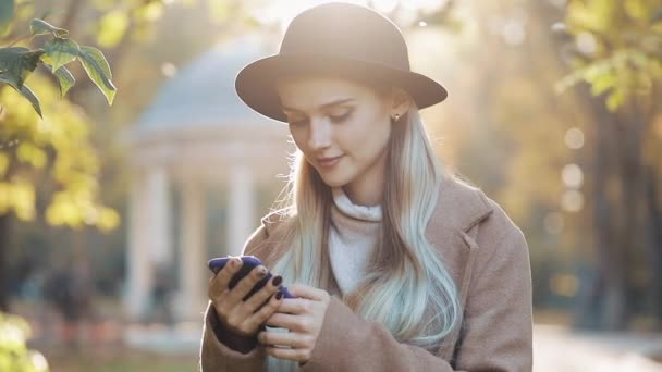 Mujer joven con un abrigo usando un teléfono inteligente de pie en el parque de otoño. Chica mirando a la cámara. Tecnología al aire libre. Luz solar — Vídeos de Stock