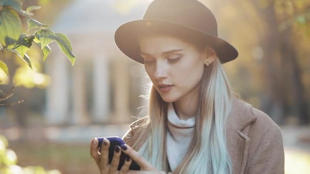 Mujer joven con un abrigo usando un teléfono inteligente de pie en el parque de otoño. Tecnología al aire libre. Luz solar — Vídeo de stock