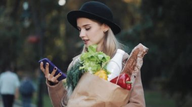 Şık ceket ayakta paket ürünlerin tutarak ve akıllı telefon ses tanıma, kullanma parkta giyen genç güzel kadın belirler düşünceler, arama mesaj