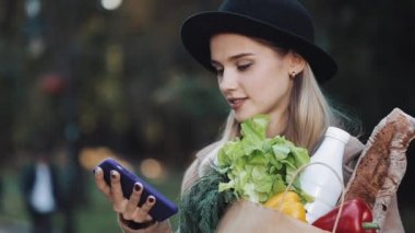Şık ceket ayakta paket ürünlerin tutarak ve akıllı telefon ses tanıma, kullanma parkta giyen genç güzel kadın belirler düşünceler, arama mesaj