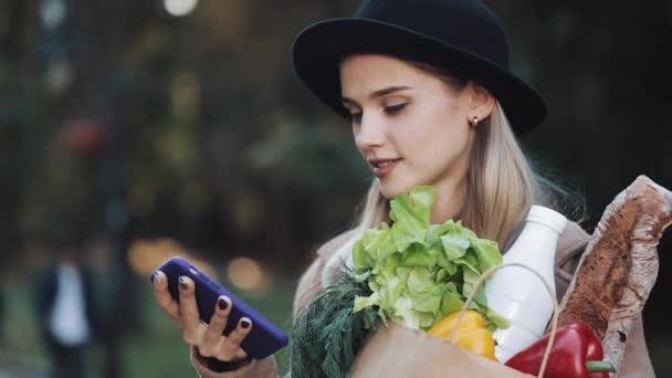 Wanita cantik muda mengenakan mantel bergaya berdiri di taman memegang paket produk dan menggunakan pengenalan suara smartphone, menentukan pikiran, pesan panggilan suara — Stok Video