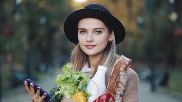 Gyönyörű fiatal nő visel álló elegáns kabát, az őszi parkban gazdaság csomag termékek és használ smartphone. Vásárlás, egészséges táplálkozás, internet-üzlet koncepció — Stock videók