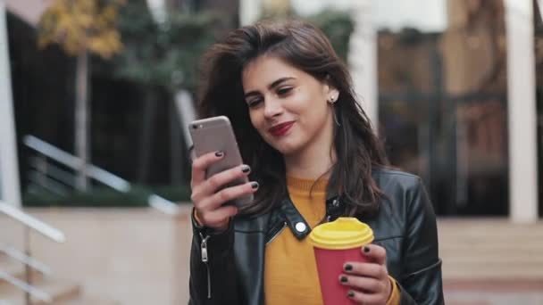 Joven mujer feliz usando teléfono inteligente y beber café para salir al aire libre, caminando por la calle de la ciudad, cámara lenta — Vídeo de stock