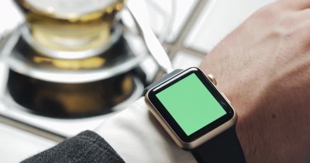 Handbewegungen aus nächster Nähe auf einer modernen Smartwatch mit grünem Bildschirm im Café. eine Tasse Tee im Hintergrund. Geschäftsmann Internet-Nachrichten-Technologie — Stockvideo