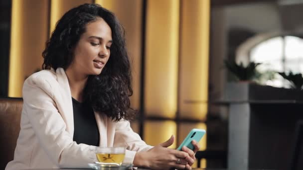 Giovane donna d'affari beve il tè in un caffè e utilizza un telefono cellulare per i social network e e-mail. Cena e navigazione web — Video Stock