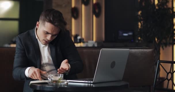 Betonade ung affärsman med laptop på kafé. Han talar med chef prata telefon och skriver anteckningar i anteckningsboken. Svårt företag, människor, tidsfristen och teknik koncept — Stockvideo