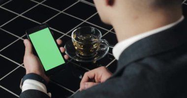 Genç işadamı kullanım smarthpone yeşil ekran dokunmatik yüzey ve el hareketi ile kafede kapatın. Hücresel, adam dokunarak ve gözatma sayfası tarafından kaydırma ve okuma Haberler