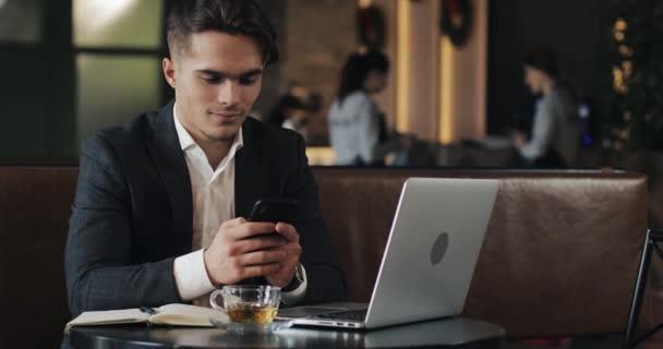 Молодий бізнесмен працює в кафе. Фрілансер використовує смартфон і ноутбук. Успішні бізнеси, ІТ, концепція фрілансера . — стокове відео