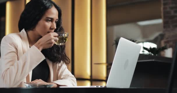 お茶を飲むと、近代的なオフィスでラップトップでビデオを見て若いビジネス女性 — ストック動画