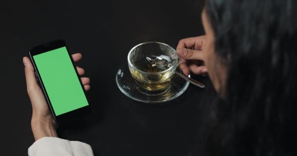 Joven mujer de negocios utilizar el teléfono inteligente con pantalla verde y beber té. Pantalla táctil en la pantalla verde del teléfono inteligente negro. Chica usando smartphone, sosteniendo smartphone con pantalla verde — Vídeo de stock