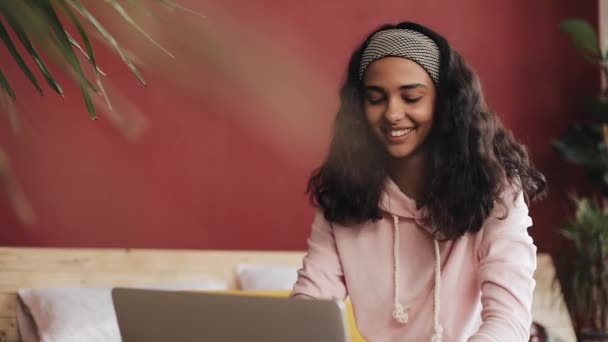 Mooi meisje gekleed in roze pyjama laptopcomputer zitten in bed thuis gebruikt. Jonge vrouw is glimlachend en chatten met vrienden — Stockvideo