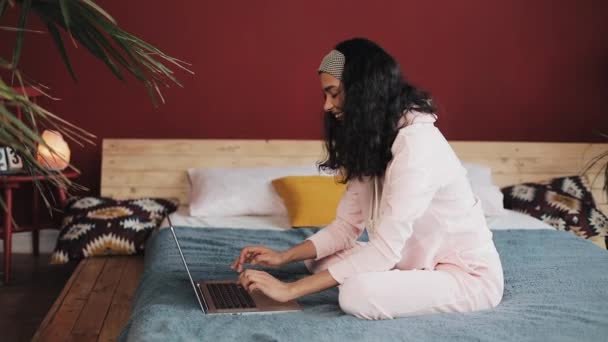 Menina bonita vestida com pijama rosa usando computador portátil sentado na cama em casa. Jovem está sorrindo e conversando com amigos — Vídeo de Stock