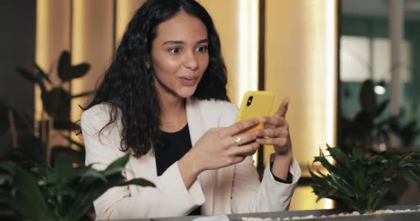Geschäftsfrau mit App auf Smartphone sitzt im modernen Büro. schöne Freizeitprofis im rosafarbenen Anzug mit guten Nachrichten auf dem Smartphone — Stockvideo