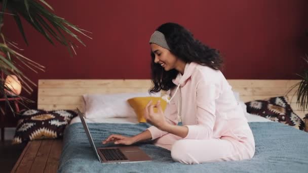 Hermosa chica vestida con pijama rosa usando computadora portátil sentada en la cama en casa. Mujer joven está sonriendo y charlando con amigos — Vídeos de Stock