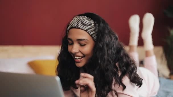 Mujer afroamericana feliz acostado en la cama usando el ordenador portátil para navegar por la web. Chica vistiendo pijama rosa sonriendo — Vídeos de Stock