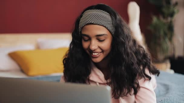 Mutlu Afrikalı-Amerikalı kadın web taramak için dizüstü bilgisayar kullanarak yatakta yatıyordu. Kız gülümseyerek Pembe pijama giyiyor — Stok video