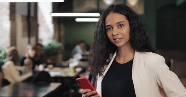 Aantrekkelijke jonge vrouw met een smartphone op zoek naar de camera glimlachen voel me gelukkig in het gezellige café — Stockvideo