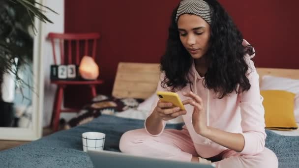 Bela menina afro-americana conversando e mensagens de texto no smartphone sentado na cama em casa. Jovem recebe boas notícias no smartphone — Vídeo de Stock