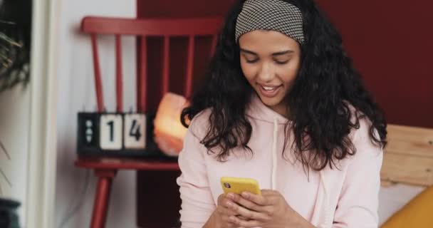 Mooie jonge vrouw die lacht tijdens het chatten op de gele smartphone zittend op het bed thuis. Meisje stuurt een bericht naar haar vriendje. Portret. Binnenkant — Stockvideo