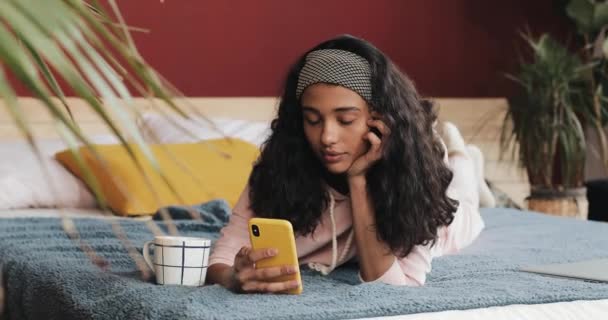 Молодая женщина смотрит видео на смартфоне лежа на кровати в спальне утром — стоковое видео