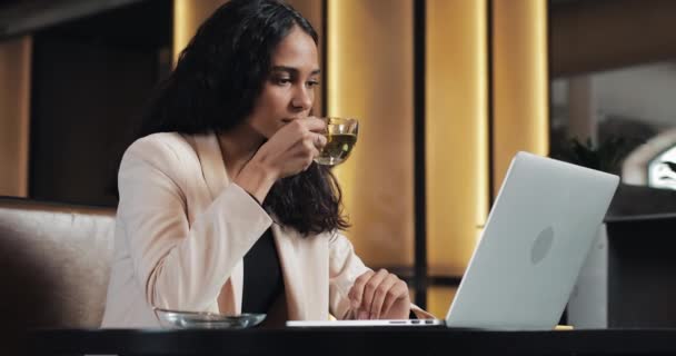 Joven mujer de negocios viendo lección en línea en el ordenador portátil. Ella sentada en la cafetería y bebiendo té — Vídeo de stock