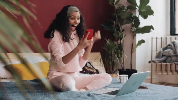 幸せなアフリカ系アメリカ人女の子チャットとテキスト メッセージングを自宅のベッドの上に座ってのスマート フォン 若い女性は スマート フォンと面白いダンスの良いニュースを受信します — ストック動画