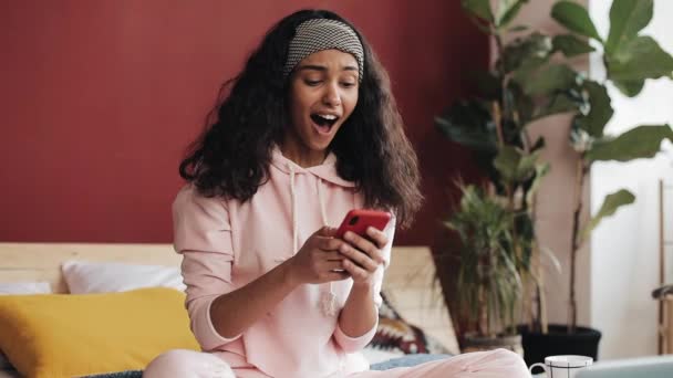 Fröhliche afrikanisch-amerikanische Mädchen im Chat und SMS-Nachrichten auf dem Smartphone sitzt auf dem Bett zu Hause. Junge Frau erhält gute Nachrichten mit Smartphone und witzigem Tanz — Stockvideo