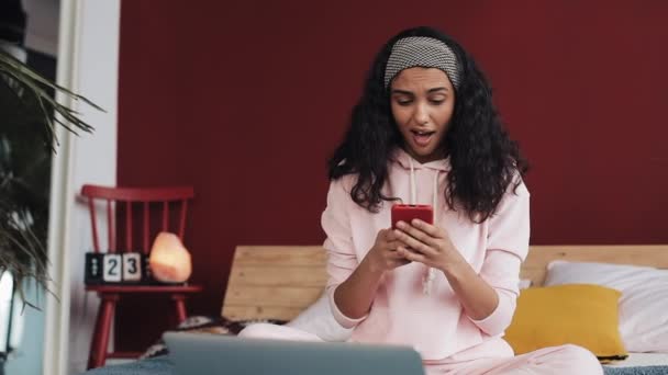 Chica afroamericana feliz charlando y mensajería de texto en el teléfono inteligente sentado en la cama en casa. Mujer joven recibe buenas noticias en el teléfono inteligente y el baile divertido — Vídeo de stock
