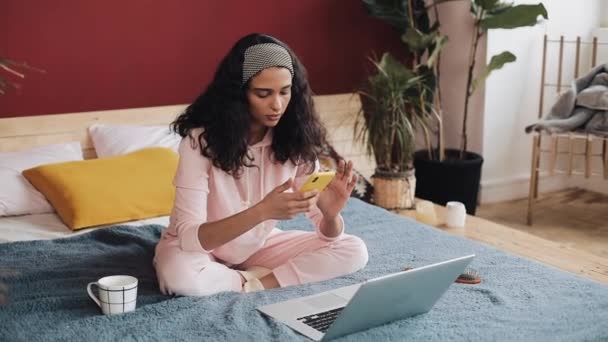 Chica afroamericana feliz charlando y mensajería de texto en el teléfono inteligente sentado en la cama en casa. Mujer joven recibe buenas noticias en el teléfono inteligente — Vídeo de stock