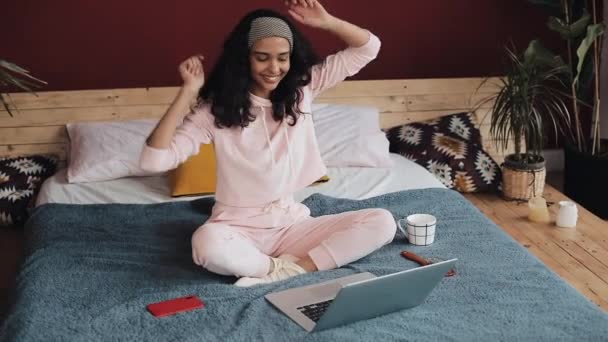 Gadis muda yang menarik bersenang-senang duduk di tempat tidur dengan laptop. Dia bernyanyi, menari, bersukacita di kabar baik — Stok Video