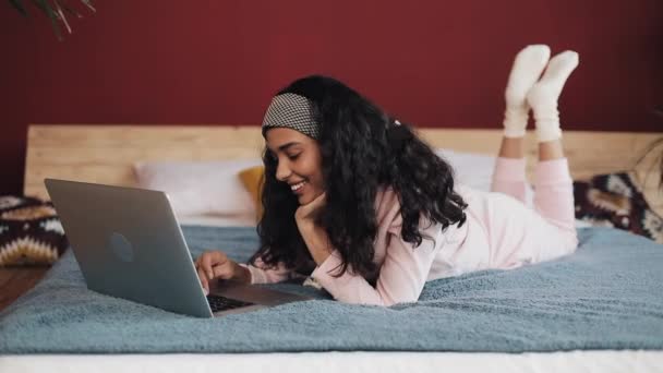 Mujer afroamericana feliz acostado en la cama usando el ordenador portátil para navegar por la web. Chica vistiendo pijama rosa sonriendo — Vídeo de stock