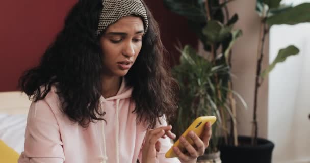 Незадоволена молода дівчина пише повідомлення на смартфоні, сидячи на ліжку вдома — стокове відео
