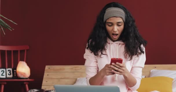 Chica afroamericana feliz charlando y mensajería de texto en el teléfono inteligente sentado en la cama en casa. Mujer joven recibe buenas noticias en el teléfono inteligente — Vídeos de Stock
