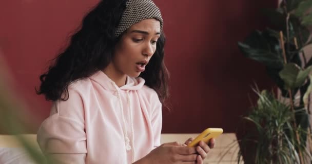 Krásná mladá žena v růžovém pyžamku vyjadřuje emoce rozhořčené otevřenými ústy s smartphone, sedí na posteli doma — Stock video