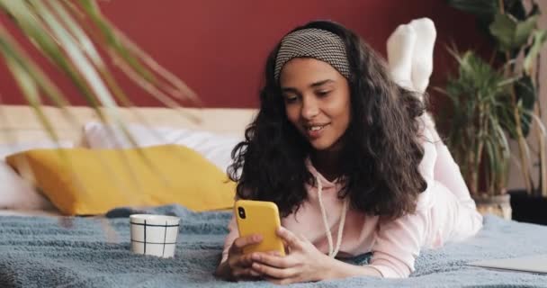 Feliz afroamericana chica se encuentra en su vientre en la cama felizmente charlando con sus amigos en una videollamada en su teléfono inteligente amarillo — Vídeo de stock