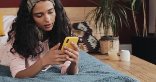 Chica afroamericana feliz charlando y mensajes de texto en el teléfono inteligente acostado en la cama en casa. Mujer joven recibe buenas noticias en el teléfono inteligente — Vídeo de stock