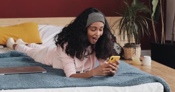 幸せなアフリカ系アメリカ人女の子チャットとテキスト ・ メッセージングを自宅のベッドに横になっているスマート フォン。若い女性は、スマート フォンの良いニュースを受信します。 — ストック動画
