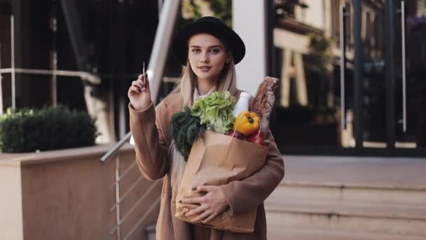 Jovem mulher bonita vestindo casaco elegante em pé na rua segurando um cartão de crédito e pacote de produtos. Menina sorrindo e olhando para a câmera. Compras, alimentação saudável — Vídeo de Stock
