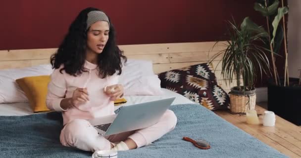 Menina atraente jovem se divertindo sentado na cama com laptop. Ela canta, dança, se alegra com as boas notícias — Vídeo de Stock