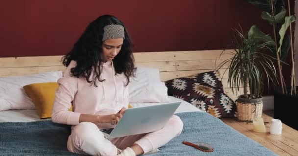 Счастливая красивая девушка в розовой пижаме работает на ноутбуке, сидя на кровати в доме. Она улыбается. — стоковое видео