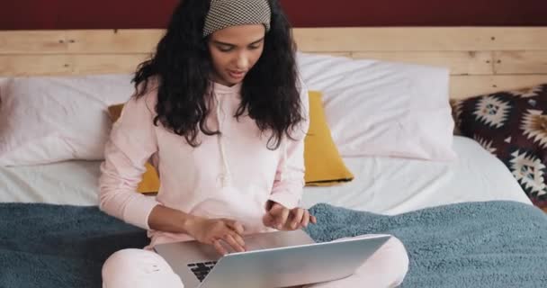 Boldog szép lány rózsaszín pizsama működő-ra egy laptop, ül az ágyon a fehér házban. Ő mosolyog