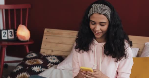 Chica afroamericana feliz charlando y mensajería de texto en el teléfono inteligente sentado en la cama en casa. Mujer joven recibe buenas noticias en el teléfono inteligente — Vídeo de stock