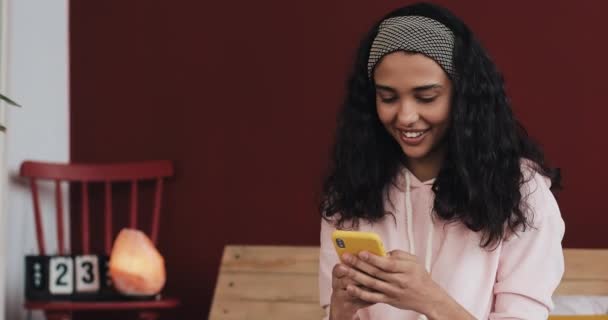 Glad afroamerikanska tjej chattar och SMS på smartphone sitter på sängen hemma. Ung kvinna får bra nyheter på smartphone — Stockvideo