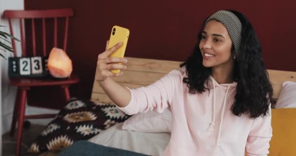 Menina americana africana feliz sentada na cama em homec hatting com seus amigos em uma chamada de vídeo em seu smartphone amarelo — Vídeo de Stock