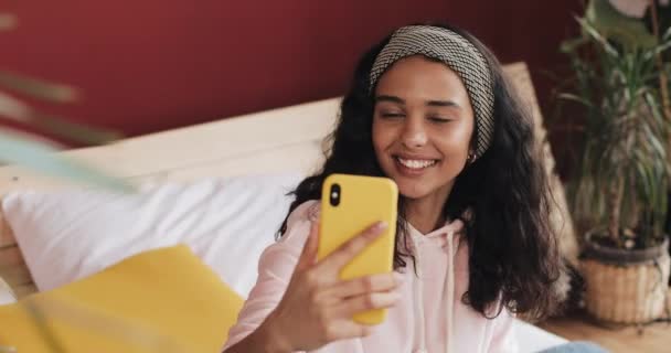 Menina americana africana feliz sentada na cama em homec hatting com seus amigos em uma chamada de vídeo em seu smartphone amarelo — Vídeo de Stock