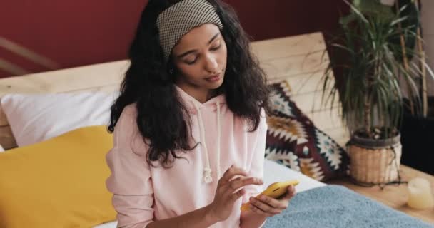 Chica afroamericana feliz charlando y mensajería de texto en el teléfono inteligente sentado en la cama en casa. Mujer joven recibe buenas noticias en el teléfono inteligente — Vídeos de Stock